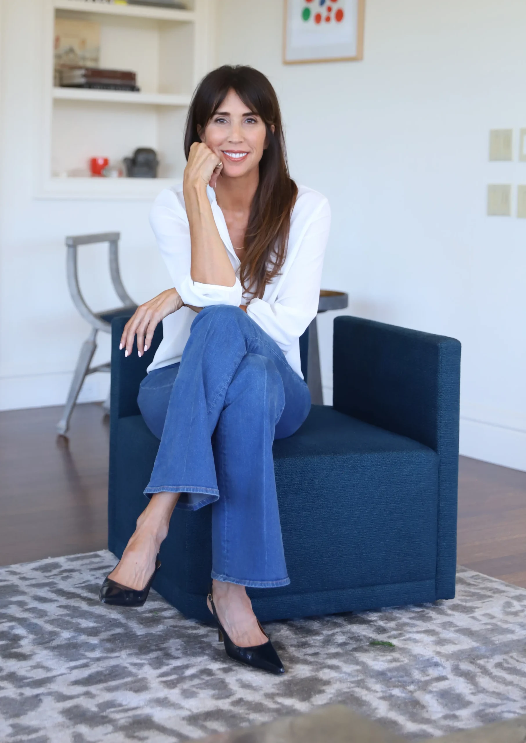 Victoria Hitchcock sitting in a blue chair with legs crossed and hand on chin, smiling at the camera wearing jeans and a white shirt